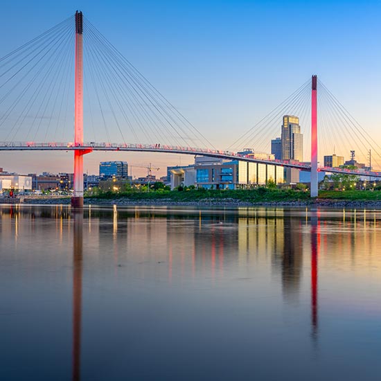 Washington Update: Banking Can Bridge the Political Divide; A suspension bridge with glowing red lights spans a calm river at dusk, reflecting in the water. City skyline with tall buildings under a clear blue sky.