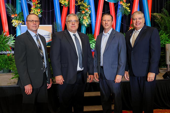 Four men in suits stand together in front of a colorful, decorated backdrop with red and blue ribbons and greenery, conveying a formal and celebratory tone.