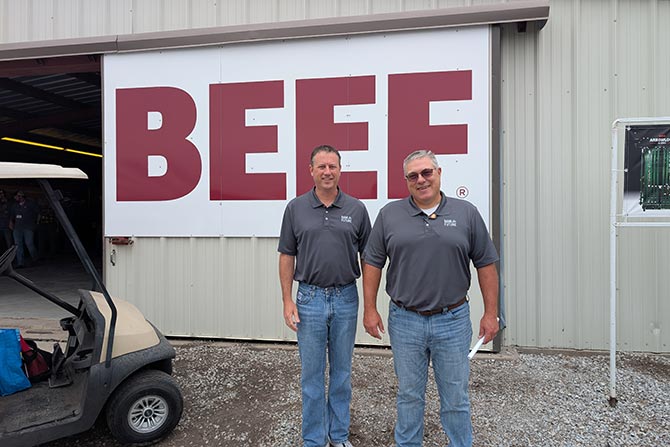 Two men in matching gray polo shirts and jeans stand smiling in front of a large building sign that reads "BEEF" in bold red letters. A golf cart is partially visible on the left.
