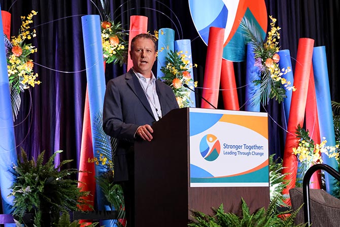 A man in a suit speaks at a podium with a "Stronger Together: Leading Through Change" sign. Colorful decor and flowers adorn the vibrant backdrop.