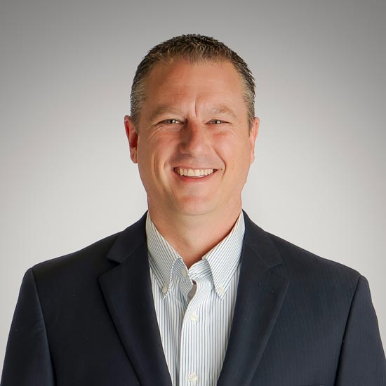 Meet Travis Sears; Smiling man in a suit with a collared shirt. He stands against a neutral gray background. The image conveys a professional and friendly tone.